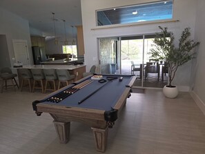 Ein Billardtisch in einem modernen Wohnzimmer mit einer Küche im Hintergrund, einem Essbereich mit Stühlen und einer großen Kübelpflanze.
