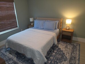 A bedroom with a large bed, two bedside tables, a nightstand, a window with blinds, and a patterned rug. - Remodeled Cape Coral Retreat with Pool, Massive Kitchen, & Entertainment Space (Cape Coral)