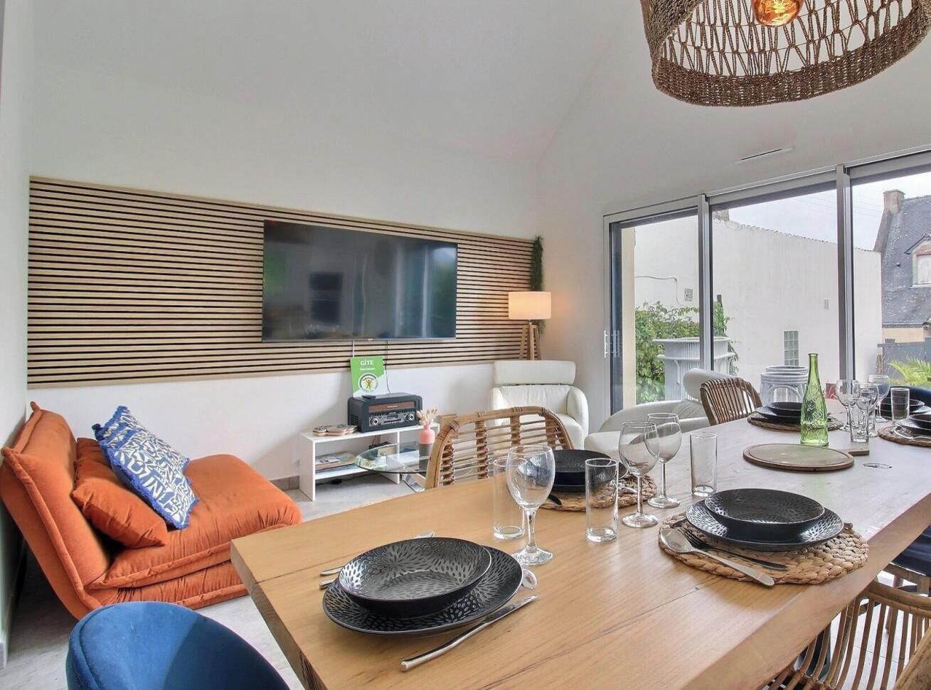 Un comedor moderno con una mesa de madera dispuesta para una comida, un sofá con cojines naranja y un televisor montado en la pared.