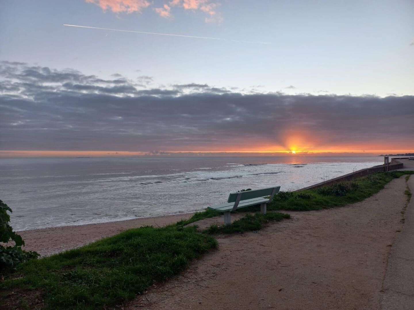 Vista de un atardecer costero con un banco y un sendero bordeando la playa.