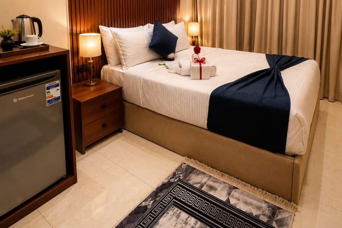 Una habitación de hotel con cama, mesitas de noche, un escritorio con hervidor, un refrigerador y una alfombra estampada.