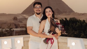 Una pareja parada frente a un gran letrero iluminado que dice 'NY', con un paisaje desértico al fondo.