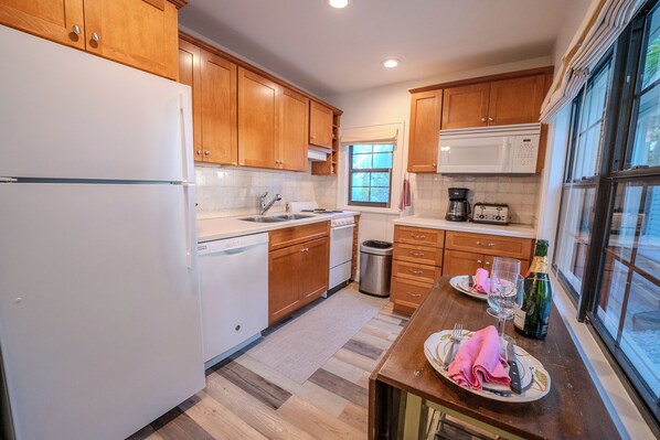 A kitchen with wooden cabinets, a white refrigerator, a microwave, and a dining table set with plates, glasses, and a bottle. - On the Beach…Retreat on the Sand—2 Units upstairs and downstairs, Sleeps 6 (Indian Rocks Beach)
