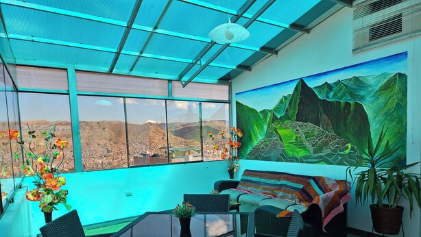 Ein Zimmer mit Glastisch, Korbmöbeln, einem großen Fenster mit Stadtblick und einer Bergwandmalerei.