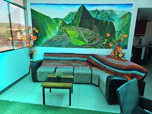 Ein Wohnzimmer mit einer Couch, einem Couchtisch, einer gemusterten Decke und einer großflächigen Wandmalerei einer Berglandschaft.