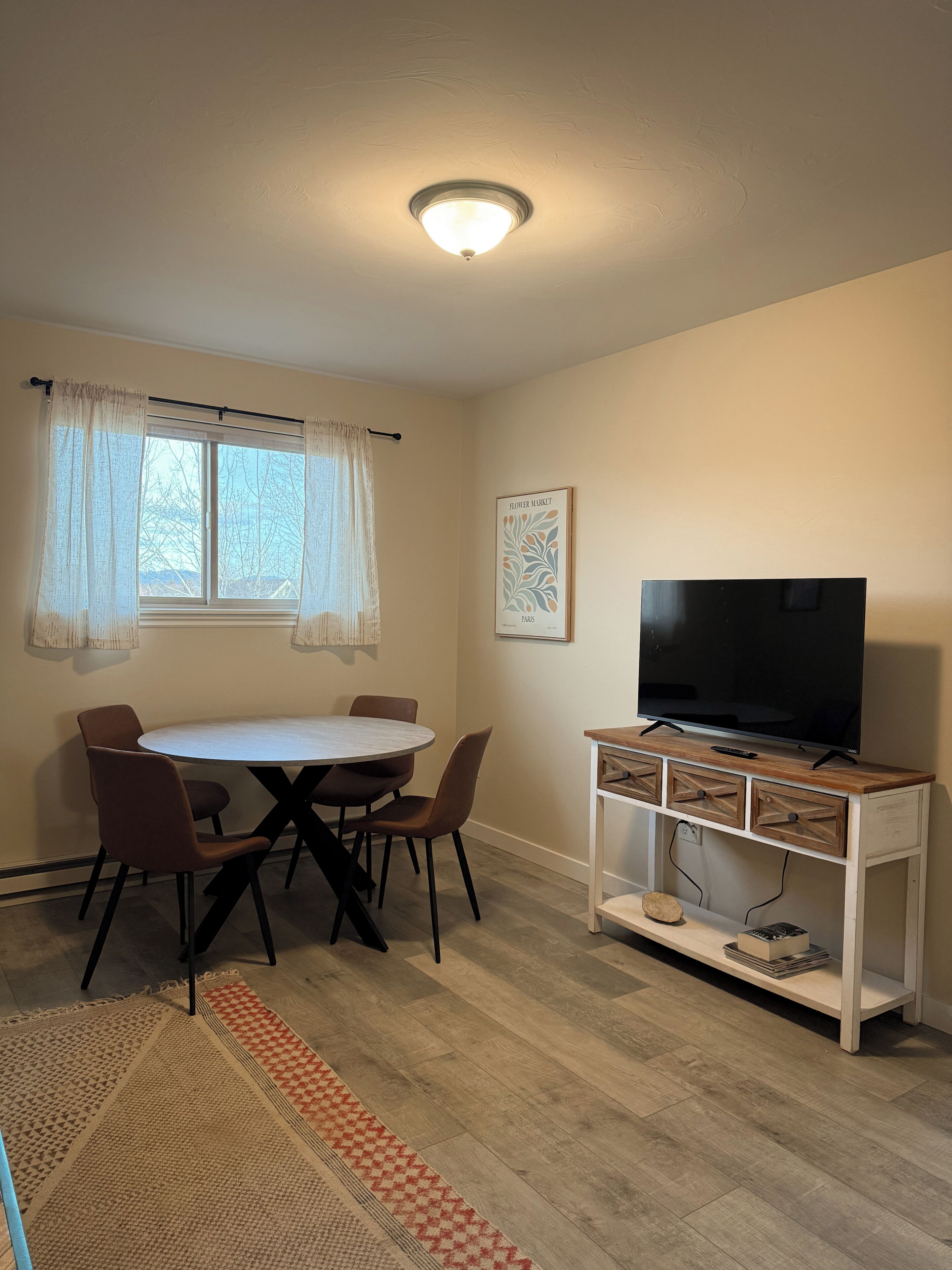 Una habitación pequeña y bien iluminada con una mesa redonda de comedor, cuatro sillas, un televisor sobre un mueble de madera y una ventana con cortinas traslúcidas.