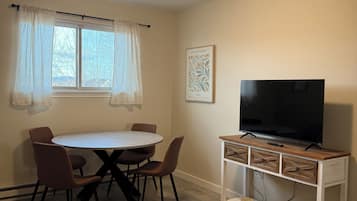 Una habitación pequeña y bien iluminada con una mesa redonda de comedor, cuatro sillas, un televisor sobre un mueble de madera y una ventana con cortinas traslúcidas.