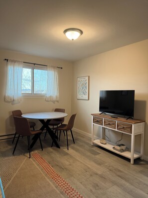 A small, well-lit room with a round dining table, four chairs, a television on a wooden stand, and a window with sheer curtains. - Bozeman Getaway Guesthouse - Suite C (Bozeman)