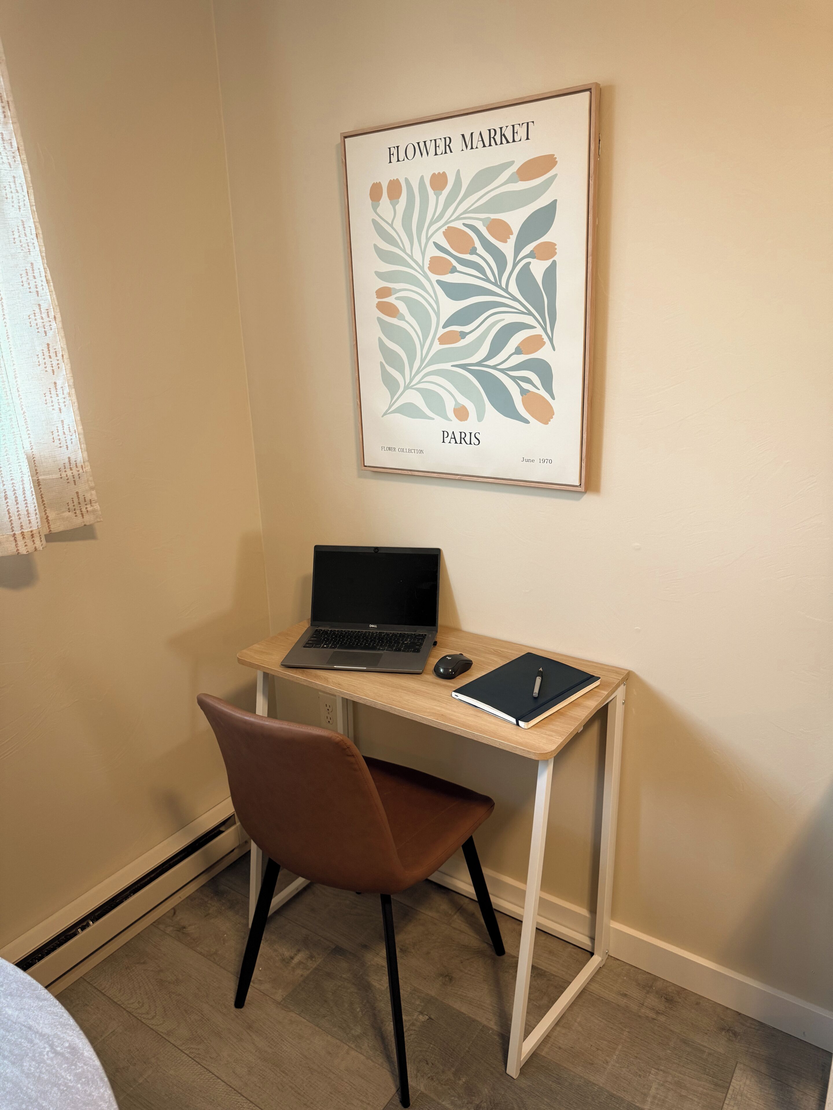 Un espacio de trabajo con una computadora portátil, un cuaderno y un bolígrafo sobre un escritorio de madera, una silla marrón y un cuadro con una imagen de un mercado de flores en la pared.