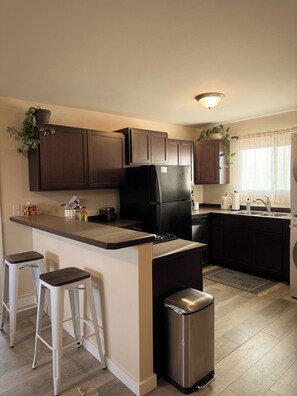 A kitchen with dark cabinets, a black refrigerator, a stainless steel trash bin, and two white bar stools. - Bozeman Getaway Guesthouse - Suite C (Bozeman)