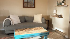A living room with a gray sofa, a wooden coffee table, a patterned rug, a wall-mounted air conditioner, a framed picture, and a shelf with decorative items. - Bozeman Getaway Guesthouse - Suite C (Bozeman)