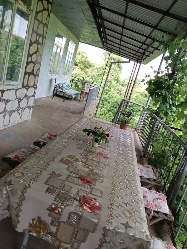 A covered balcony with a table set with a tablecloth and flowers, a chair, and a potted plant.