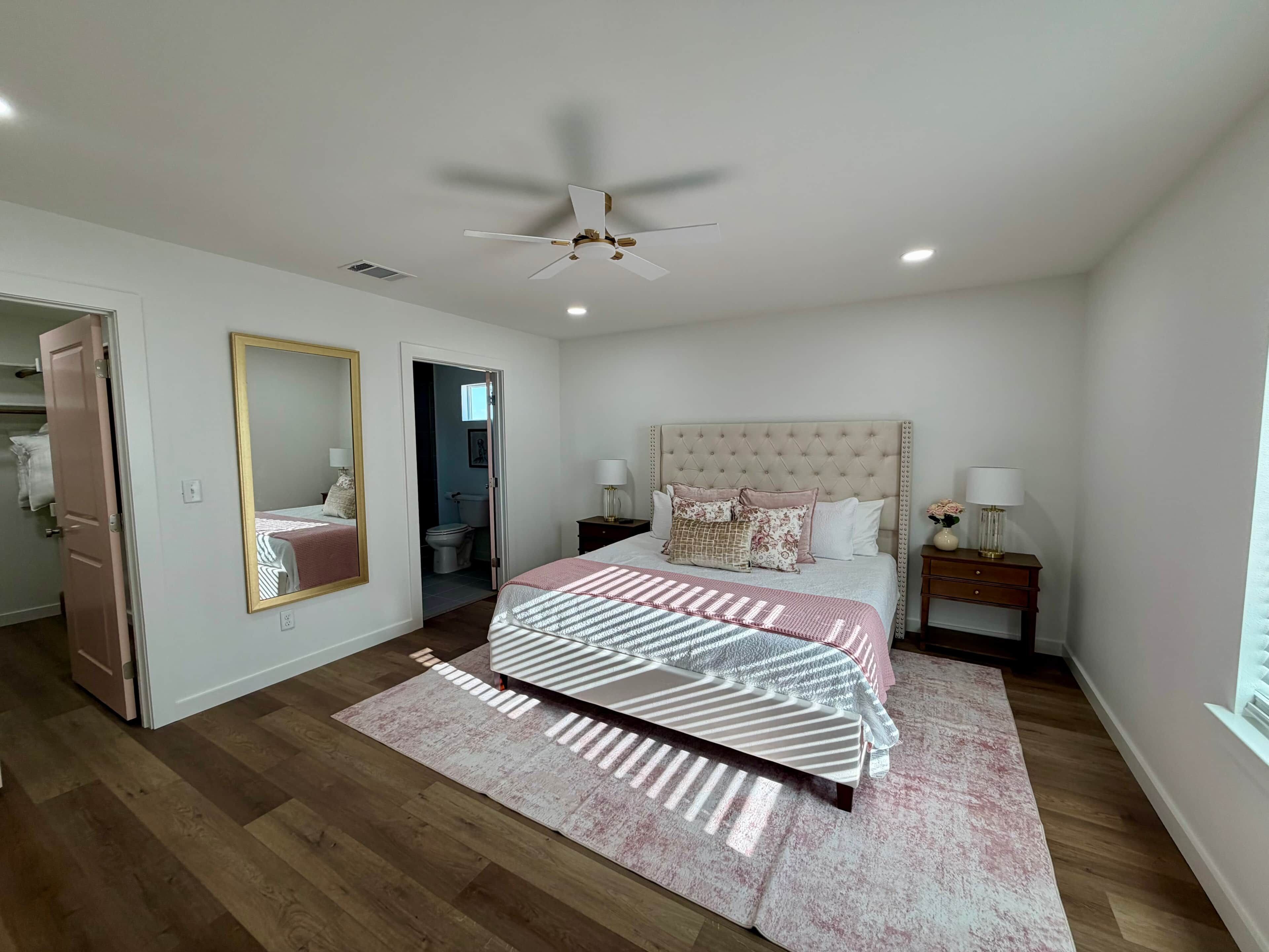 A bedroom with a bed, a ceiling fan, a nightstand, a mirror, and a door leading to another room.