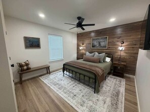 A bedroom with a bed, a bench, a ceiling fan, and wall-mounted lights. - Hunter's Lodge by WinStar - King Bed (Love County)