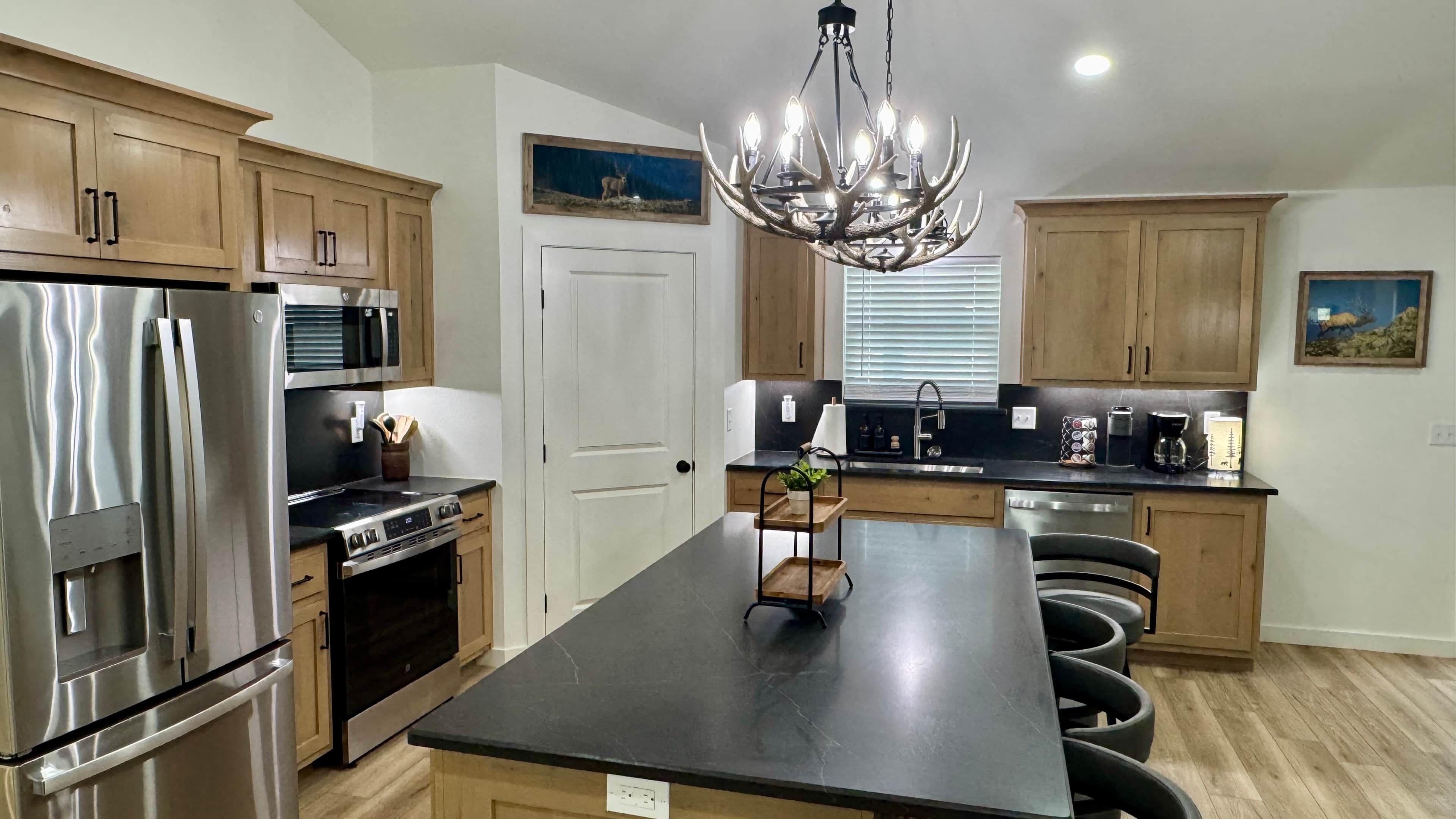 A modern kitchen with stainless steel appliances, wooden cabinets, a central island with bar stools, and a chandelier.