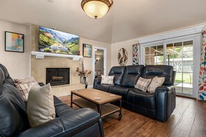 A living room with a fireplace, a black leather sofa, a wooden coffee table, and a large flat-screen TV mounted above the fireplace. - Cheerful Oasis w/ Private Hot Tub, Yard, & Big Rooms (Fort Worth)
