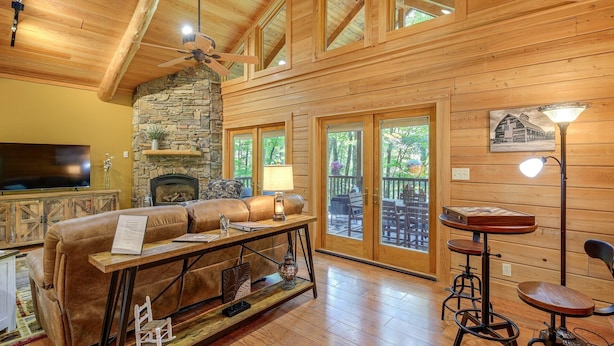 Un intérieur de chalet chaleureux avec une cheminée en pierre, un canapé en cuir, une table à manger en bois et un balcon doté de meubles d’extérieur.