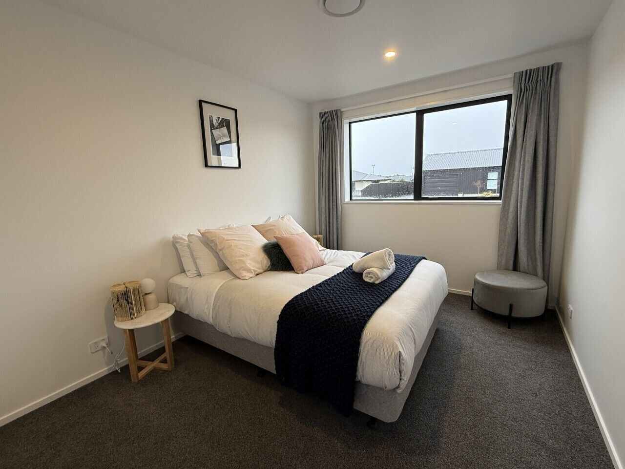A bedroom with a bed, a small side table, a round ottoman, a window with curtains, and a framed picture on the wall.