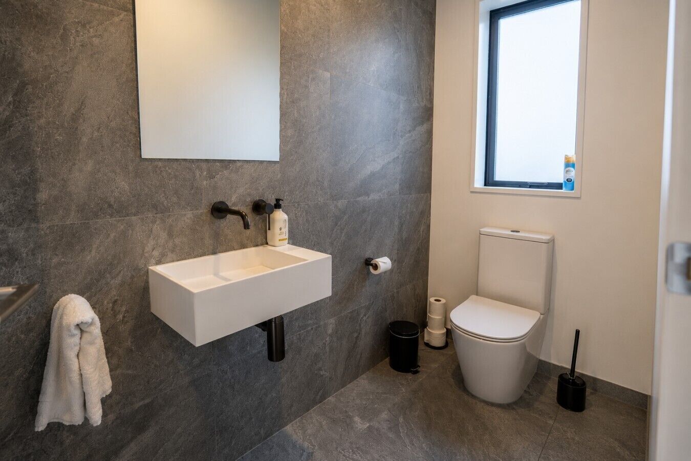 A bathroom with a white sink, dark floor tiles, a toilet, and a window.