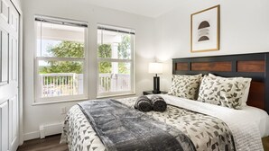 Ein Schlafzimmer mit einem Bett, zwei Fenstern, einem Nachttisch, einer Lampe und einem gerahmten Bild an der Wand.