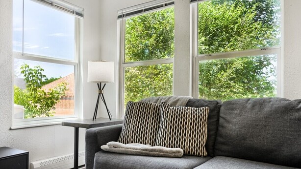 Ein modernes Wohnzimmer mit einem grauen Sofa, gemusterten Kissen, einer Stehlampe und großen Fenstern mit Blick auf Bäume und ein Ziegelgebäude.
