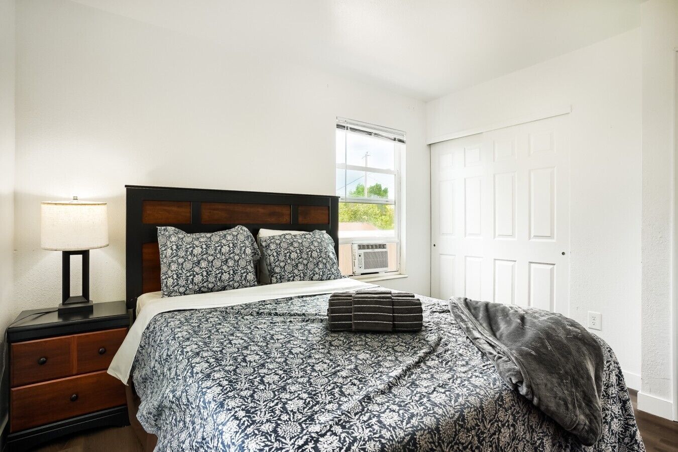 A bedroom with a bed, a nightstand, a lamp, a window, and a door.