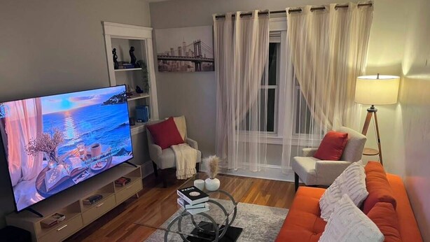 Un salon avec un téléviseur, un canapé, des fauteuils, une lampe et une fenêtre ornée de rideaux légers.