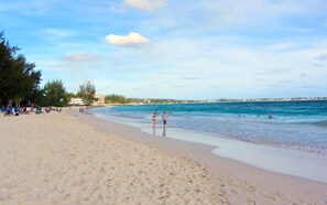 Ein Strand mit klarem, blauem Wasser, Menschen, die schwimmen und am Ufer entlanggehen, sowie eine Reihe von Liegestühlen und Sonnenschirmen auf der linken Seite.