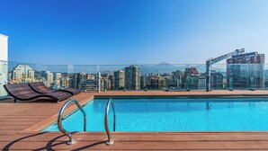 A rooftop pool with lounge chairs, a clear view of a cityscape, and modern buildings in the background. - Nest Collection El Golf (Las Condes)
