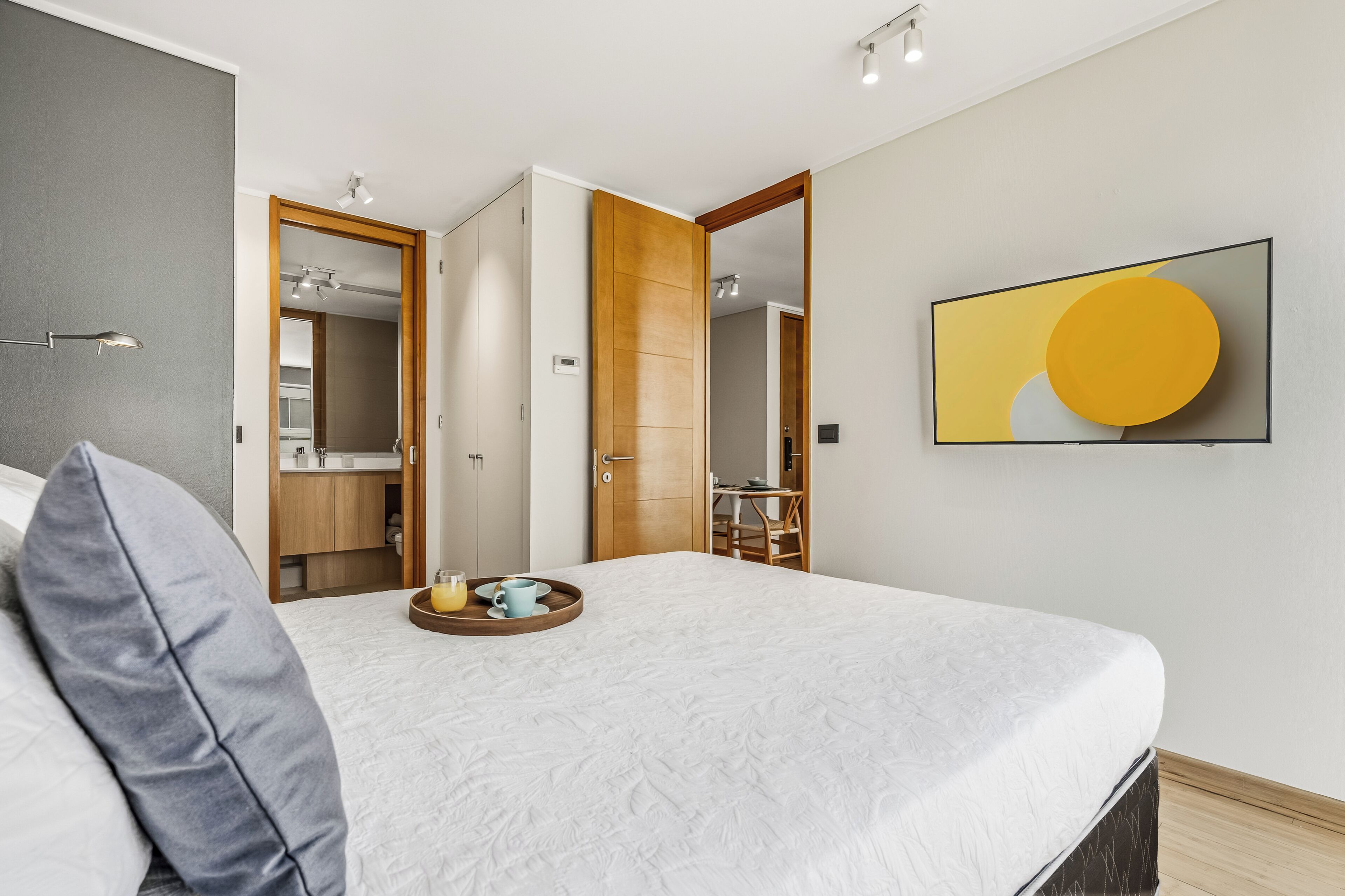 A bedroom with a bed, a bedside table with a tray, a mirror, a wooden door, and a painting on the wall.