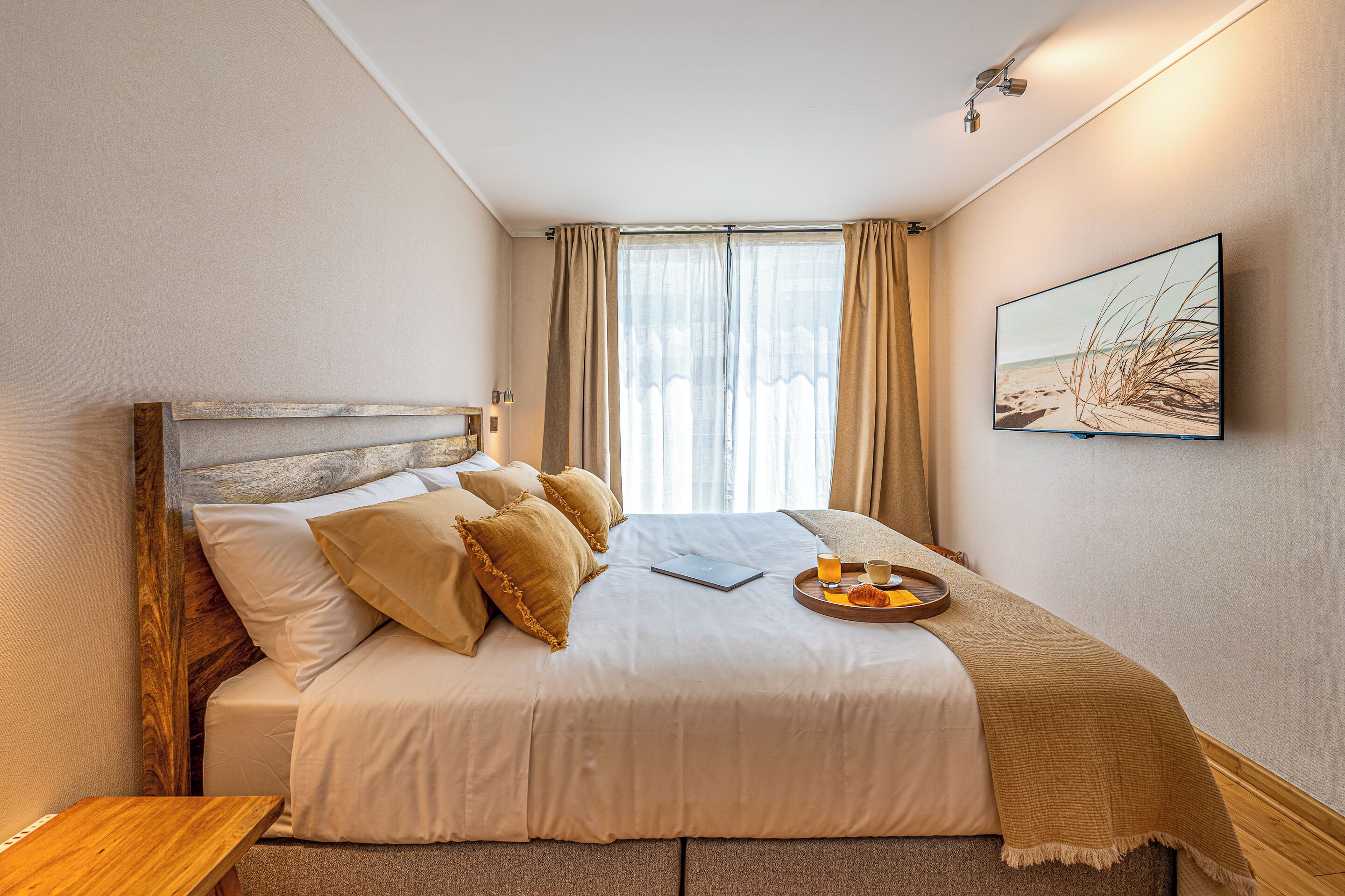 A neatly made bed with pillows, a bedside table, a tray with a drink, and a beach-themed picture on the wall.