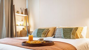 A neatly made bed with a tray of pastries and a glass of orange juice, a bedside table with a vase and candles, and a shelf with decorative items. - Nest Collection El Golf (Las Condes)