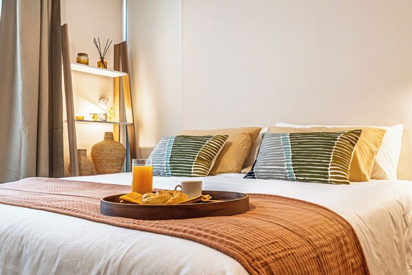 Una cama bien hecha con una bandeja de pasteles y un vaso de jugo de naranja, una mesita de noche con un florero y velas, y una estantería con objetos decorativos.