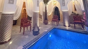 Un área de piscina interior lujosa con arquitectura ornamentada, incluyendo arcos y columnas, y una piscina con un borde de mosaico azul.