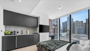 Une chambre moderne avec un grand lit, une vue sur la ville, une cuisine aux meubles sombres et un téléviseur à écran plat.