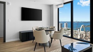 Un espace de repas moderne avec une table ronde, des chaises et une grande fenêtre donnant sur un paysage urbain et l’eau.