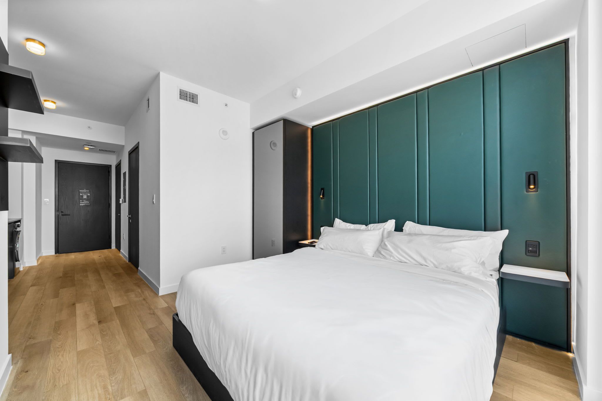 Quarto de hotel moderno com uma cama grande, piso de madeira, parede de destaque azul-esverdeada e banheiro separado.