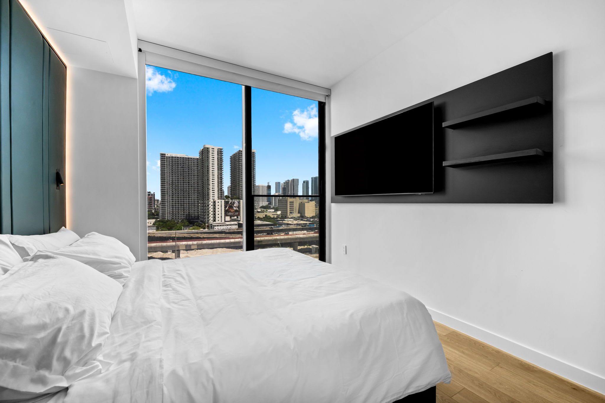 Un dormitorio moderno con una cama grande, un televisor montado en la pared y vista a la ciudad a través de un ventanal.