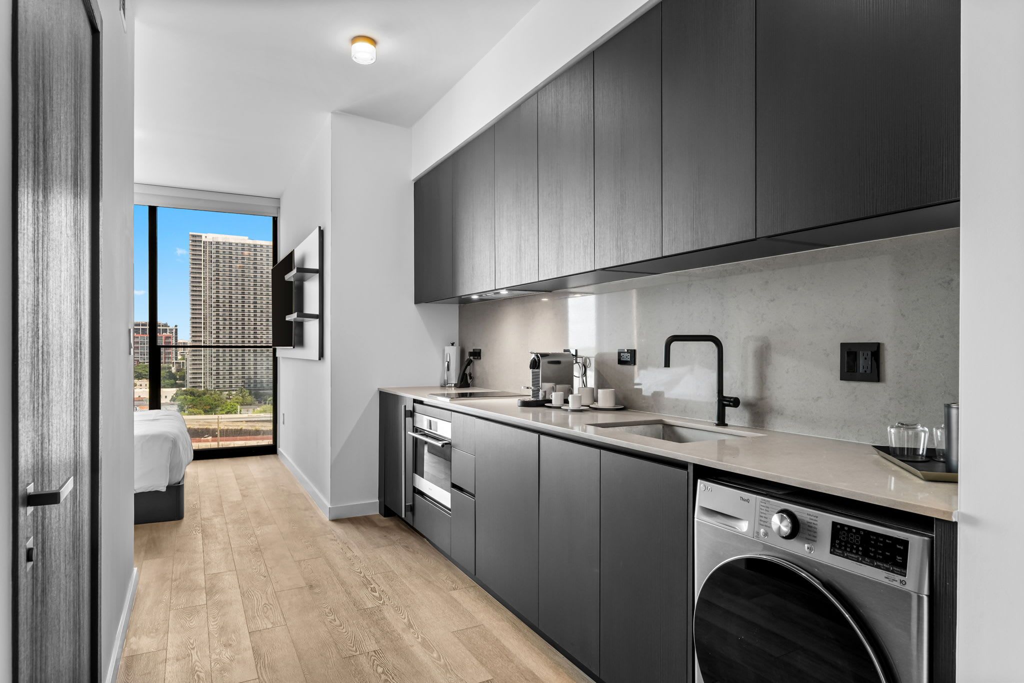 Een moderne keuken met grijze kastjes, een inbouwoven, een vaatwasser en een groot raam met uitzicht op een stadsbeeld.