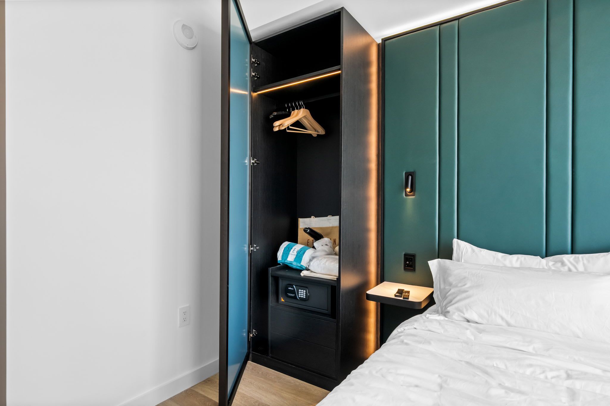 Une chambre d’hôtel comprenant un lit, une armoire avec une tringle pour suspendre les vêtements, une table de chevet avec une lampe et un panneau de commande.