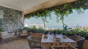 Un balcon avec une table à manger pour quatre personnes, entouré de chaises en osier, d’un canapé et offrant une vue sur la ville et la mer.
