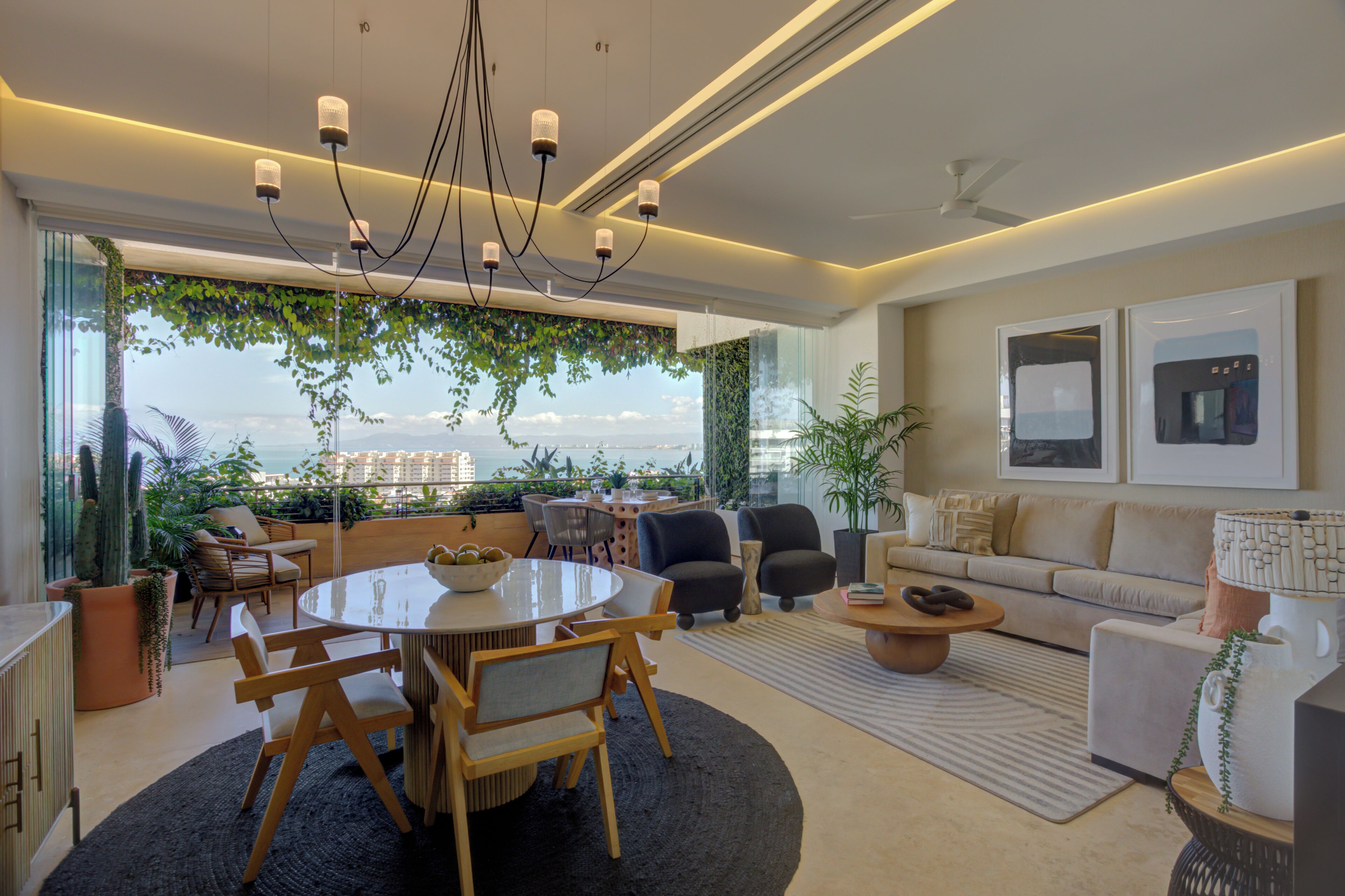 A modern living room with a round dining table, sofas, and chairs. There is a large window with a view of the city and greenery.