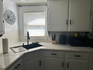 Una cocina con armarios blancos, un grifo negro, una ventana con persianas y un microondas en la encimera.