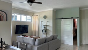 A living room with a grey sofa, a TV, a ceiling fan, and a dining area with a table and chairs. - Spacious, cool, and tastefully decorated one bedroom with pull out sofa. (Kingston)