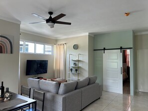 Ein Wohnzimmer mit grauem Sofa, einem Fernseher, einem Deckenventilator und einem Essbereich mit Tisch und Stühlen.
