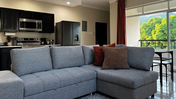 A modern living room with a grey sectional sofa, a kitchen area with a microwave, stove, and refrigerator, and a balcony with a view of greenery. - Spacious, cool, and tastefully decorated one bedroom with pull out sofa. (Kingston)