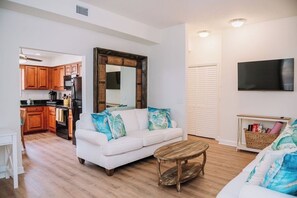 A living room with a white sofa, wooden coffee table, and a kitchen area with wooden cabinets and appliances. - Stay Downtown St. Augustine 3BR and Parking (St. Augustine)