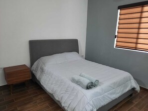 Un lit simple avec une tête de lit grise, une table de chevet en bois et une fenêtre avec des stores horizontaux.