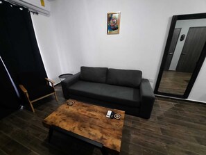 Un salon avec un canapé noir, une table basse en bois, une petite table d’appoint et un tableau accroché au mur.