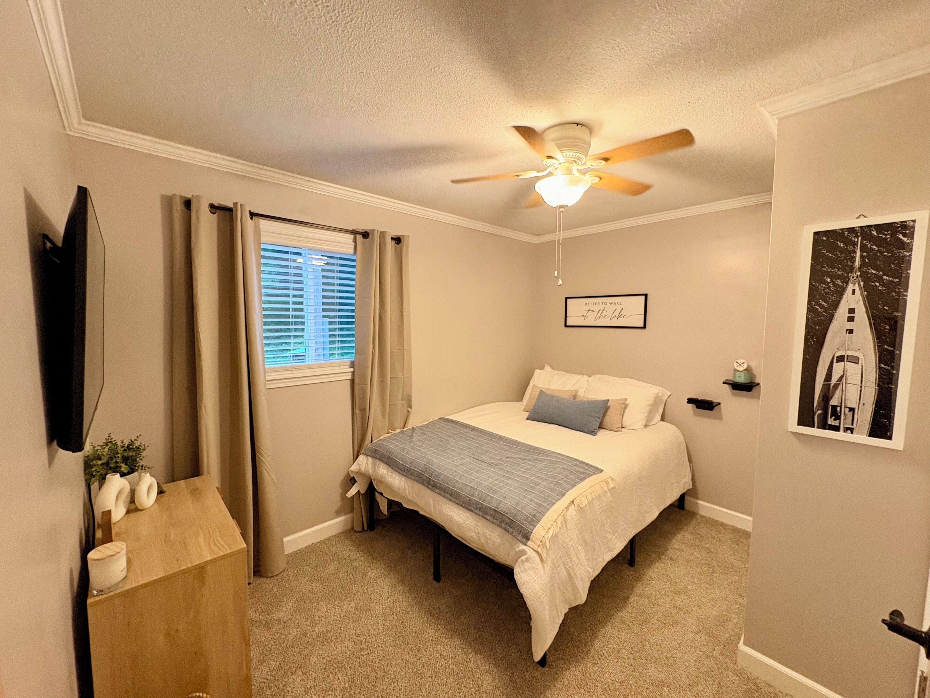 Une chambre à coucher avec un lit, un ventilateur de plafond, une fenêtre avec des rideaux, un téléviseur fixé au mur, un tableau encadré et une petite étagère.